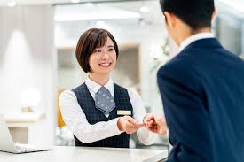 Hotel front desk where a Japanese staff member hands a calligraphy box labeled Japanese Calligraphy to a traveler