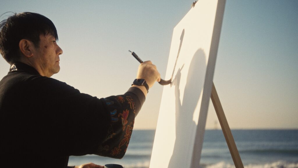 Artist Madara creating Hisseki-jutsu calligraphy on a large canvas by the sea