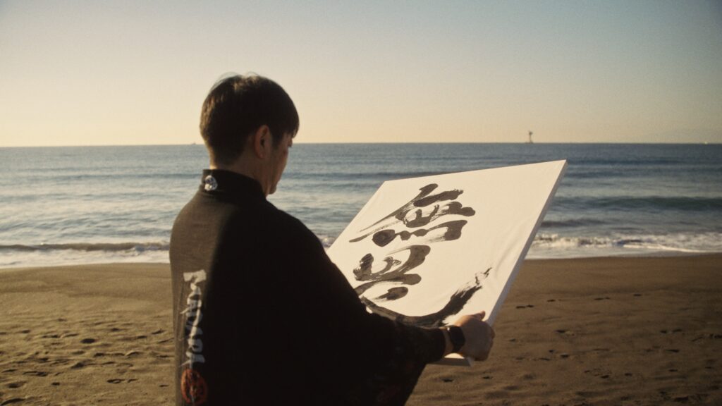 Madara holding a finished calligraphy artwork while standing quietly by the sea.