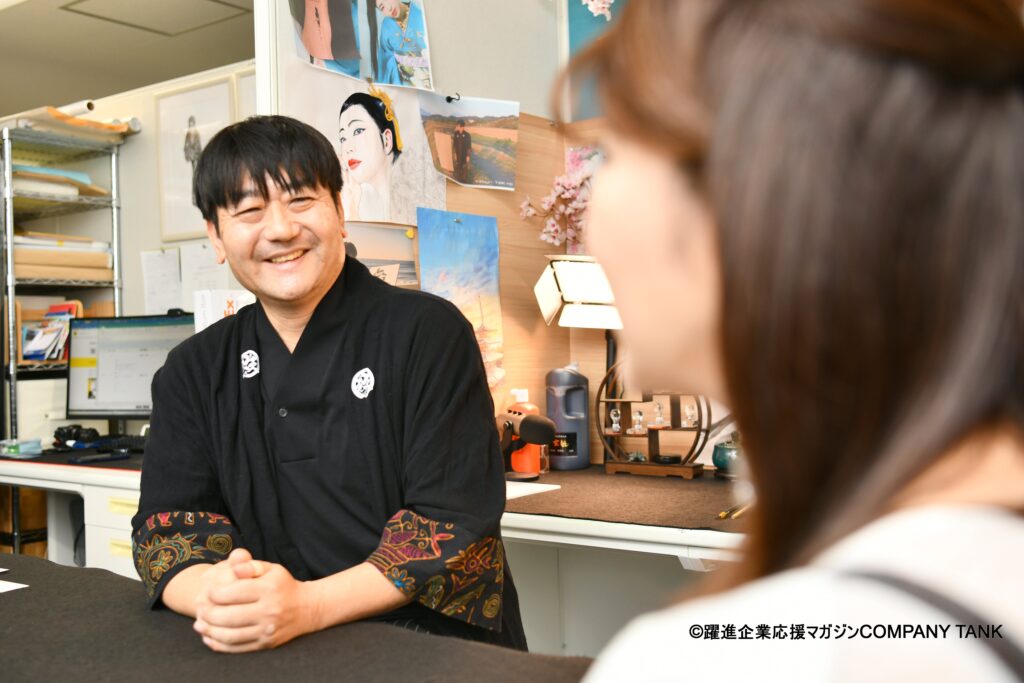 Japanese calligraphy artist speaking with an interviewer during a magazine feature at the KANJI TOKYO studio, surrounded by artworks and tools.