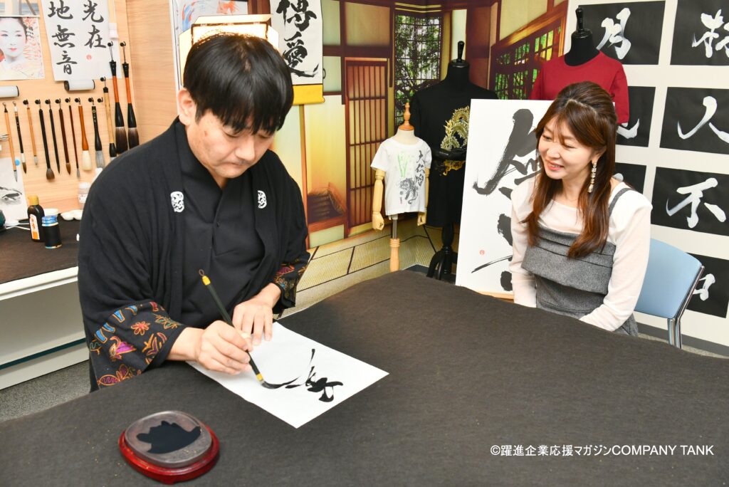 Japanese calligraphy artist MADARA demonstrating Hisseki-jutsu during a magazine interview, observed by TV personality Miho Yabe at the KANJI TOKYO studio.