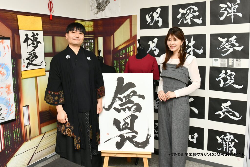 Japanese calligraphy artist MADARA standing with TV personality Miho Yabe during a COMPANY TANK magazine interview, showcasing a large Hisseki-jutsu calligraphy work at the KANJI TOKYO studio.