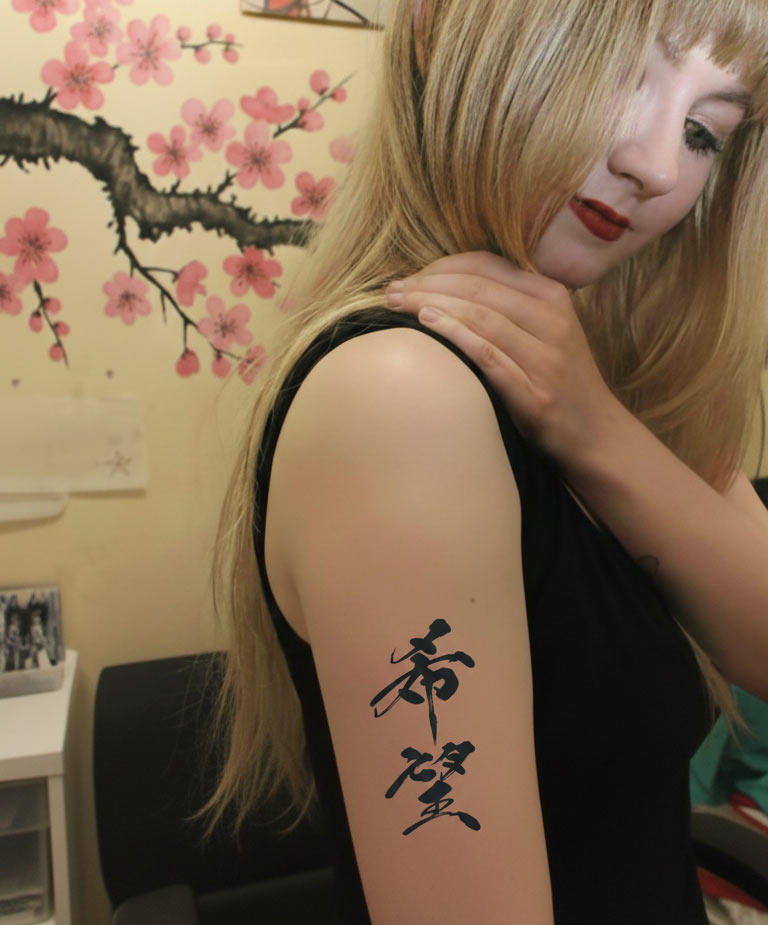 A blonde woman in a black top posing in a studio, touching her right shoulder. A small, blueish-black tattoo of the Japanese kanji "Hope" (希望), styled in traditional brush calligraphy, is visible on her upper arm. The background features cherry blossom art and a pop-art style poster.