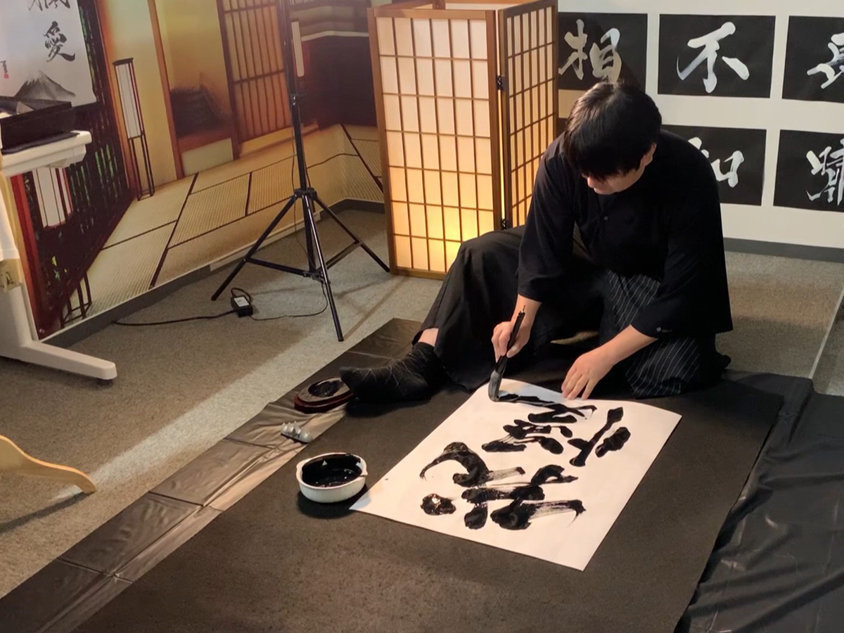 Calligraphy artist MADARA creating the kanji “Gaman” (endurance) for a custom tattoo design at KANJI TOKYO, carefully writing each stroke by hand during the production process.