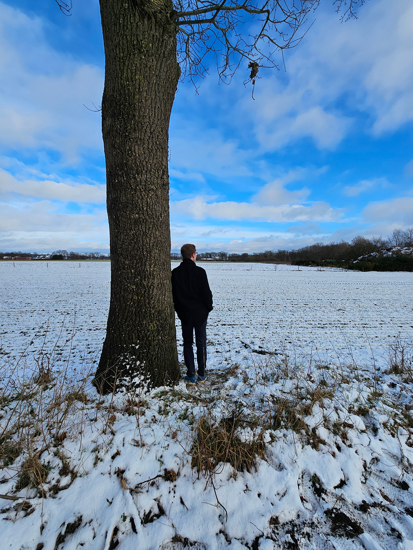A man standing alone beside a large tree in a snow-covered field, quietly reflecting on life and his inner thoughts