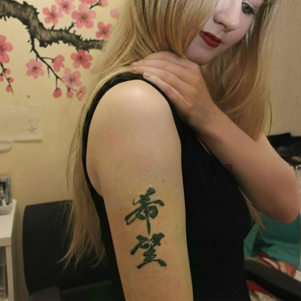 A blonde woman in a black top posing in a studio, touching her right shoulder. A small, blueish-black tattoo of the Japanese kanji "Hope" (希望), styled in traditional brush calligraphy, is visible on her upper arm. The background features cherry blossom art and a pop-art style poster.